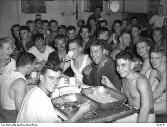 CREW MEMBERS ENJOYING A MEAL, SOME EATING FROM THE LARGE BAKING PANS, IN HMAS SYDNEY STOKERS MESS.