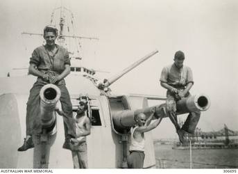 ALEXANDRIA, EGYPT. 1940-07-21. MEMBERS OF THE SHIP'S COMPANY, BOB LINKLATER (LEFT) AND JACK CLARK, SITTING ON THE 6 INCH GUN BARRELS OF A TURRET OF THE CRUISER HMAS SYDNEY (II) AS THEY PREPARE THEM FOR REPAINTING AFTER THE PAINT HAD BEEN DAMAGED DURING THE ACTION OFF CAPE SPADA. TWO FLEET RESERVE RATINGS ARE WORKING ON THE BARRELS IN THE BACKGROUND. SIMILAR WORK IS BEING CARRIED OUT ON B TURRET WHICH IS TRAINED TO PORT WITH THE RIGHT GUN ELEVATED. (NAVAL HISTORICAL COLLECTION).