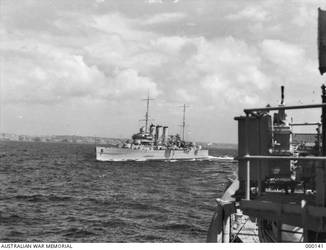OFF SYDNEY, NSW. PORT BOW VIEW OF THE HEAVY CRUISER HMAS AUSTRALIA (II) AS SHE APPEARED AFTER HER 1938-1939 REFIT. THE SHIP HAS HAD HER BRIDGEWORK EXTENDED. THE FOUR SINGLE 4 INCH MK V AA GUNS HAVE BEEN REPLACED BY EIGHT TWIN 4 INCH MK XVI WEAPONS ON MK XIX MOUNTINGS. THE OLD 4 INCH GUNDECK HAS BEEN REMOVED AND THE NEW MOUNTINGS PLACED ON THE UPPER DECK. A NEW DIRECTOR CONTROL TOWER HAS BEEN FITTED AND THE ORIGINAL MOVED AFT, REPLACING THE HIGH ANGLE CONTROL POSITION AND THE ORIGINAL CRANE. A SEARCHLIGHT TOWER HAS BEEN ADDED JUST BEFORE IT. HIGH ANGLE CONTROL POSITIONS HAVE BEEN ADDED IN THE BRIDGE WINGS. NEW CRANES HAVE BEEN ADDED AMIDSHIPS, PORT AND STARBOARD. THE FOUR SINGLE TWO POUNDER AA GUNS HAVE BEEN REPLACED BY QUADRUPLE 2 POUNDER MOUNTINGS ON PLATFORMS PORT AND STARBOARD JUST FORWARD OF THE SEARCHLIGHT TOWER. HER MAIN ARMAMENT AND DIRECTOR ARE TRAINED TO PORT. THE SEAGULL V AMPHIBIAN IS ON ITS CATAPULT. (NAVAL HISTORICAL COLLECTION).