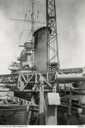 ALEXANDRIA, EGYPT. 1940-07-19. DETAIL VIEW OF THE CRUISER HMAS SYDNEY (II) AMIDSHIPS, LOOKING FORWARD FROM THE PORT 4 INCH GUN DECK. IN THE FOREGROUND IS THE SHIP'S CATAPULT AND AIRCRAFT HANDLING CRANE, BEHIND WHICH IS THE FORWARD FUNNEL SHOWING THE HIT MADE BY A 6 INCH SHELL FROM THE ITALIAN CRUISER BARTOLOMEO COLLEONI IN THE ACTION OFF CAPE SPADA. (NAVAL HISTORICAL COLLECTION).