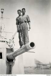 ALEXANDRIA, EGYPT. 1940-07-21. TWO MEMBERS OF THE SHIP'S COMPANY, BOB LINKLATER AND JACK CLARK, STANDING ON LEFT 6 INCH GUN OF A TURRET ABOARD THE CRUISER HMAS SYDNEY (II). THE COAL WHARF AND DERRICK EQUIPMENT ARE IN THE RIGHT BACKGROUND. (NAVAL HISTORICAL COLLECTION).