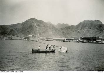 ADEN. 1940-05. NATIVE BOATMEN SECURING THE BOW LINE OF THE CRUISER HMAS SYDNEY (II). THE TOWNSHIP AND SURROUNDING MOUNTAINS ARE IN THE BACKGROUND. (NAVAL HISTORICAL COLLECTION).