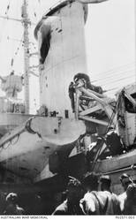 Unidentified Australian sailors inspecting the damage caused to the light cruiser HMAS Sydney II, after the port side of the forward funnel was struck by a shell fired from the Italian cruiser Bartolomeo Colleoni during a naval battle off Cape Spada, Crete. The Italian ship was immobilised by accurate fire from the Sydney's six inch guns and then sunk by torpedo's launched from the escorting British destroyers. Also visible, is part of the crane (right) used to recover the Seagull V (Walrus) amphibian aircraft.