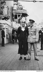 Informal portrait of 23464 Able Seaman (AB) Martin Curtis James (left), of Newcastle, NSW, and 35338 Leading Aircraftman (LAC) Keith Homard, 9 Squadron RAAF, of Broadmeadow, Newcastle, NSW, on the starboard deck of HMAS Sydney II. Both men were amongst the 645 crew members on board HMAS Sydney lost on 19 November 1941 when the Sydney was sunk in the Indian Ocean off the coast of Western Australia by the German auxiliary cruiser Kormoran. A despatch clerk prior to enlistment, AB James was aged 19 years of age at the time of his death. A travelling salesman prior to enlistment, LAC Homard had enlisted as a photographer in the RAAF on 10 March 1941, as he had previously had five years experience as a press photographer at the Newcastle Sun newspaper. LAC Homard was 27 years of age at the time of his death. The view of the deck looking forward shows many of the ship's features including the deck made of 7 inch wide Borneo white hardwood planks, an unidentified crew member on the ship's crane, and a 36 inch searchlight mounted on its sponson abreast the forward funnel. Lashed around the sponson are splinter mats. Behind AB James and LAC Homard are empy boat crutches which normally contain two of the ship's 35 foot motor boats, indicating that the ship is at anchor or moored to a buoy. The unidentified sub lieutenant at the left is standing at the aft end of the ship's bakery.