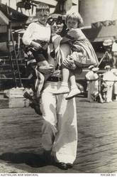 Informal portrait of 19508 Petty Officer (PO) William Lewis Aylott, HMAS Sydney, of McMahons Point, NSW, holding his children Bill and Dawn in his arms on a wharf at Circular Quay. HMAS Sydney II, in the background, had returned to Sydney the previous evening following service in the Mediterranean. PO Aylot and his son are draped in a Union Jack flag. PO Aylott has a tattoo of a butterfly on his left hand and is wearing an aluminium identification bracelet which was made on the ship. Originally from Devonport, England, he had enlisted in the RAN in 1930. PO Aylott was killed in action, aged 32, when HMAS Sydney II was sunk in the Indian Ocean by the German auxiliary cruiser Kormoran on 20 November 1941.